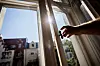 Hand öffnet ein Fenster in heller Wohnung bei sonnigem Wetter; draußen Häuser und blauer Himmel.