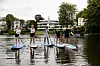 Fünf Personen paddeln stehend auf Boards über einen ruhigen Fluss vor Bäumen und Häusern.