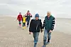 Drei Generationen spazieren am Strand, weitere Familienmitglieder folgen im Hintergrund.