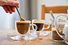 Frau rührt in einer Hamburger Wohnung Kaffee um, auf dem Tisch stehen Tassen und ein Zuckerglas.