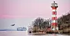 Rot-weiß gestreifter Leuchtturm an winterlicher Küste, im Hintergrund ein Frachtschiff auf dem Wasser.