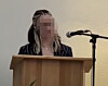 Person am Pult spricht neben einem Mikrofon vor hellem Hintergrund.