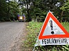 Feuerwehr-Warnschild am Straßenrand, im Hintergrund ein Einsatzfahrzeug auf einer Waldstraße.