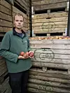 Mann in grüner Jacke hält Äpfel vor gestapelten Holzkisten mit weiteren Äpfeln.