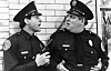 Steve Guttenberg und Peter Van Norden als Polizisten vor einer Ziegelwand im Gespräch.