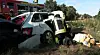 Feuerwehrkräfte an einem schwer beschädigten Auto neben einer verletzten Person im Gras an einer Straße.