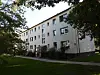 Mehrstöckiges Wohnhaus mit hellen Fassaden, Eingangsbereich und Bäumen vor einer gepflegten Rasenfläche.