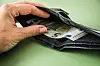 Hand öffnet schwarzes Portemonnaie mit 5-Euro-Schein auf grünem Hintergrund.