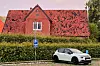 Rotes Backsteinhaus hinter Hecke, davor ein weißes SUV und ein Parkschild.