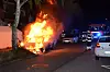 Auto brennt am Straßenrand, Feuerwehrfahrzeuge und Einsatzkräfte im Hintergrund.