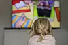 Blondes Mädchen mit Zopf liegt auf dem Sofa und schaut ein buntes Kinderprogramm im Fernsehen.