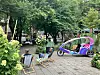 Zwei bunte Rikschas auf einem gepflasterten Platz im Park, daneben Stühle und ein Tisch unter Bäumen.