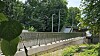 Fußgängerbrücke mit Metallgeländer über grüner Uferlandschaft, Bäume und Gebäude im Hintergrund.