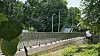 Fußgängerbrücke mit Metallgeländer über grüner Uferlandschaft, Bäume und Gebäude im Hintergrund.