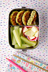 Quinoa-Brokkoli-Bratlinge in Brotdose mit Gurke und Apfel, darunter bunte Schulutensilien.