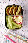 Quinoa-Brokkoli-Bratlinge in Brotdose mit Gurke und Apfel, darunter bunte Schulutensilien.