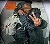 Zwei Personen vor einer Wand, eine mit heller Tasche, die andere zeigt ein Friedenszeichen.