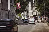 Mehrere Autos auf einer Stadtstraße, im Vordergrund ein verwischtes vorbeifahrendes Fahrzeug, links eine rot-weiße Fahne.