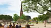 Menschen im Park, roter Kirchturm im Hintergrund, Bäume und Rasen bei sonnigem Wetter.
