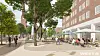 Menschen an einem Platz mit Bäumen, Radweg und Straßencafé vor einem roten Backsteingebäude.