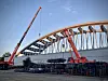 Zwei Kräne montieren eine geschwungene Brücke auf einer Baustelle.
