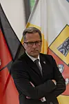 Mann im schwarzen Anzug steht mit verschränkten Armen vor Fahnen.