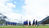 Fußballspieler trainieren auf einem Rasenplatz vor Bergen und wolkigem Himmel.