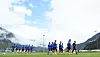 Fußballspieler trainieren auf einem Rasenplatz vor Bergen und wolkigem Himmel.
