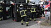 Feuerwehrleute arbeiten an einem umgestürzten Auto am Straßenrand, ein Rettungsfahrzeug steht dahinter.