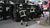 Feuerwehrleute arbeiten an einem umgestürzten Auto am Straßenrand, ein Rettungsfahrzeug steht dahinter.