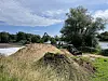 Erdhaufen und Geröll an einer Uferbaustelle mit Bäumen, Fluss und bewölktem Himmel.