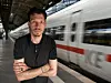 Mann am Bahnsteig vor vorbeifahrendem weißen Zug mit rotem Streifen.