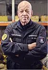 Feuerwehrmann mit verschränkten Armen vor Regalen mit Einsatzmaterial in einer Halle.