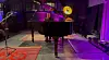 Pianist am schwarzen Flügel auf einer Bühne mit violetter Beleuchtung und Mikrofonständern.