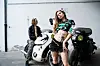 Drei junge Menschen posieren vor zwei Motorrädern an einer weißen Wand.