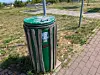 Grüner Mülleimer neben einem gepflasterten Weg auf grasigem Gelände.