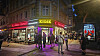 Menschen stehen vor einem beleuchteten Kiosk an einer Straßenecke bei Nacht.