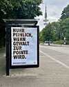 Großes Plakat mit provokantem Spruch an einer Straße, im Hintergrund ein Fernsehturm und Bäume.