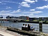 Menschen sitzen auf einer Bank an der Uferpromenade mit Blick auf Fluss und Stadt.
