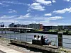Menschen sitzen auf einer Bank an der Uferpromenade mit Blick auf Fluss und Stadt.