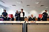 Mehrere Personen in einem Sitzungssaal an Tischen, einige sitzen, andere stehen und sprechen.