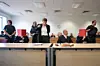 Mehrere Personen in einem Sitzungssaal an Tischen, einige sitzen, andere stehen und sprechen.