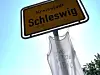 Ortsschild Schleswig über transparentem Protestbanner im Freien.