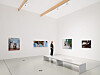 Person steht in weißem Ausstellungsraum vor mehreren gerahmten Fotografien und einer langen Sitzbank.