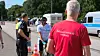Polizisten kontrollieren einen Radfahrer an einer Straße, im Vordergrund ein Mann im roten T-Shirt.