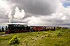 Brockenbahn mit Dampflok und Waggons im Nationalpark Harz auf einer grünen Wiese unter dunklen Wolken.
