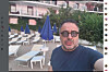 Mann im Selfie vor Poolbereich mit Liegestühlen und blauen Sonnenschirmen.