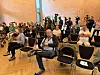 Zuschauer sitzen in einem Saal, im Hintergrund filmen Kamerateams weitere Besucher.