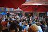 Viele Menschen sitzen und stehen unter roten Sonnenschirmen vor einem Backsteingebäude.