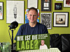 Mann mit Plakat „Wie ist die Lage?“ vor grüner Wand mit Bildern und Mikrofon.